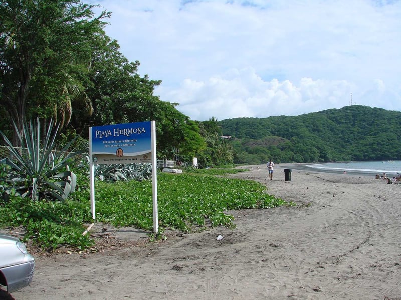 The sign at the entrance to Playa Hermosa, with the long grey sand beach curving away behind it